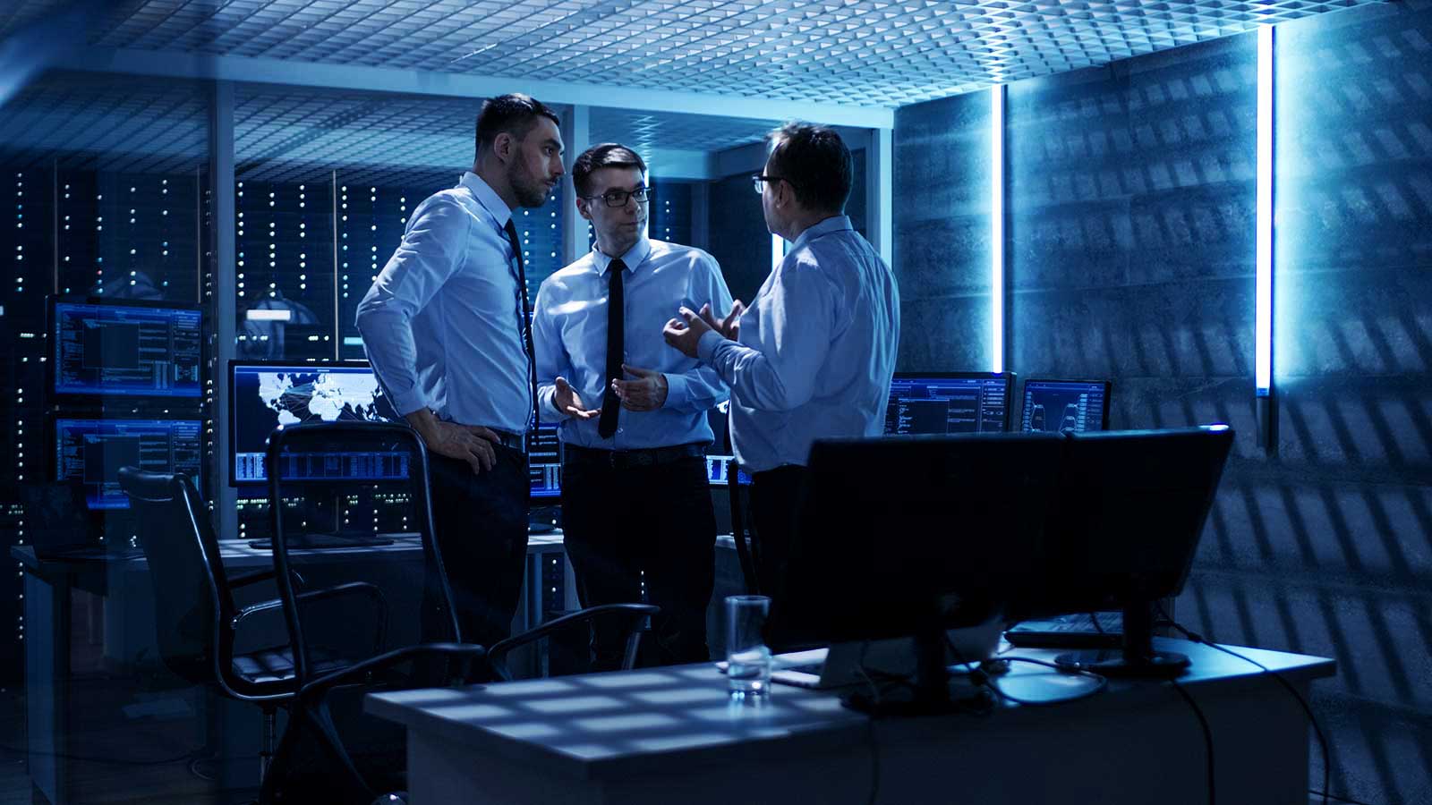 Three men in formal attire are standing and discussing something in a dimly lit, high-tech control room. The room is filled with computer monitors displaying data and graphs. The atmosphere suggests a serious and focused conversation.