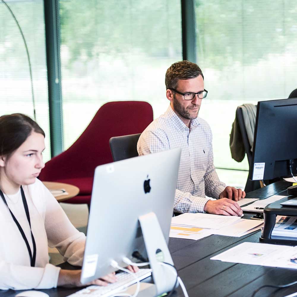 Two people working at computers in a modern office. The woman on the left is using an iMac, and the man on the right is wearing glasses and working on a desktop. Papers and office equipment are on the desk, with large windows and green scenery in the background.