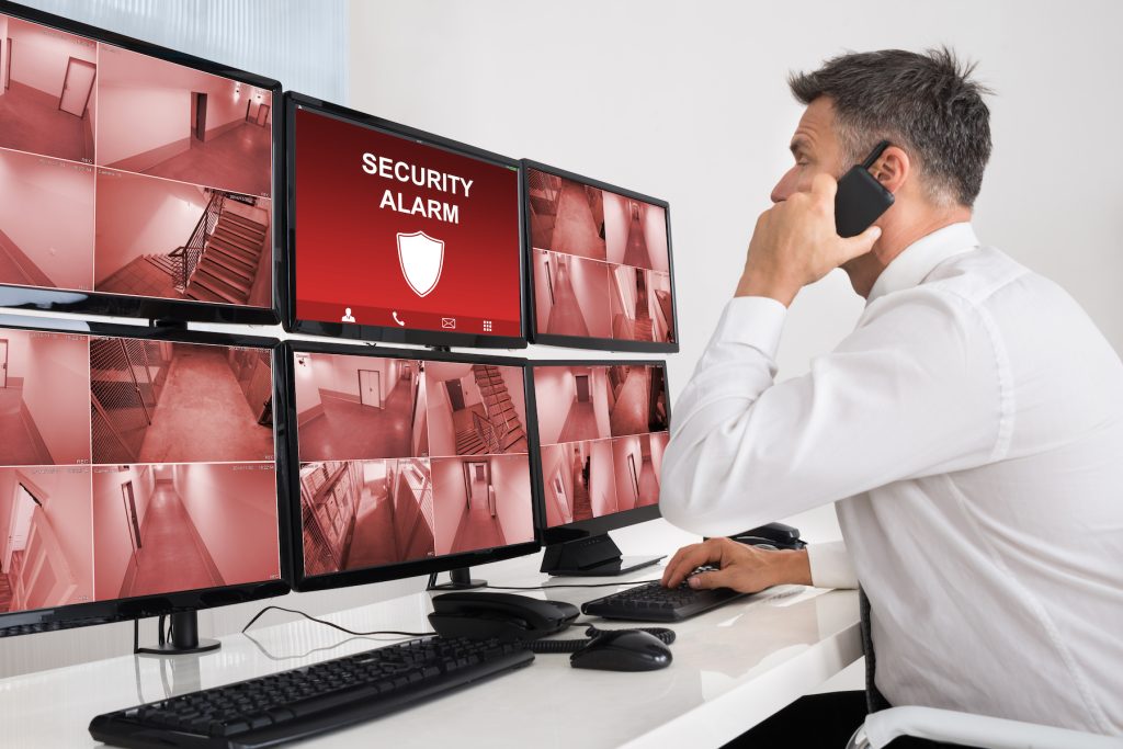 A man in a white shirt is monitoring multiple security camera feeds on several computer screens at his desk. He is speaking on a phone, likely coordinating with his Managed IT Services team, and one of the screens displays a "Security Alarm" alert.