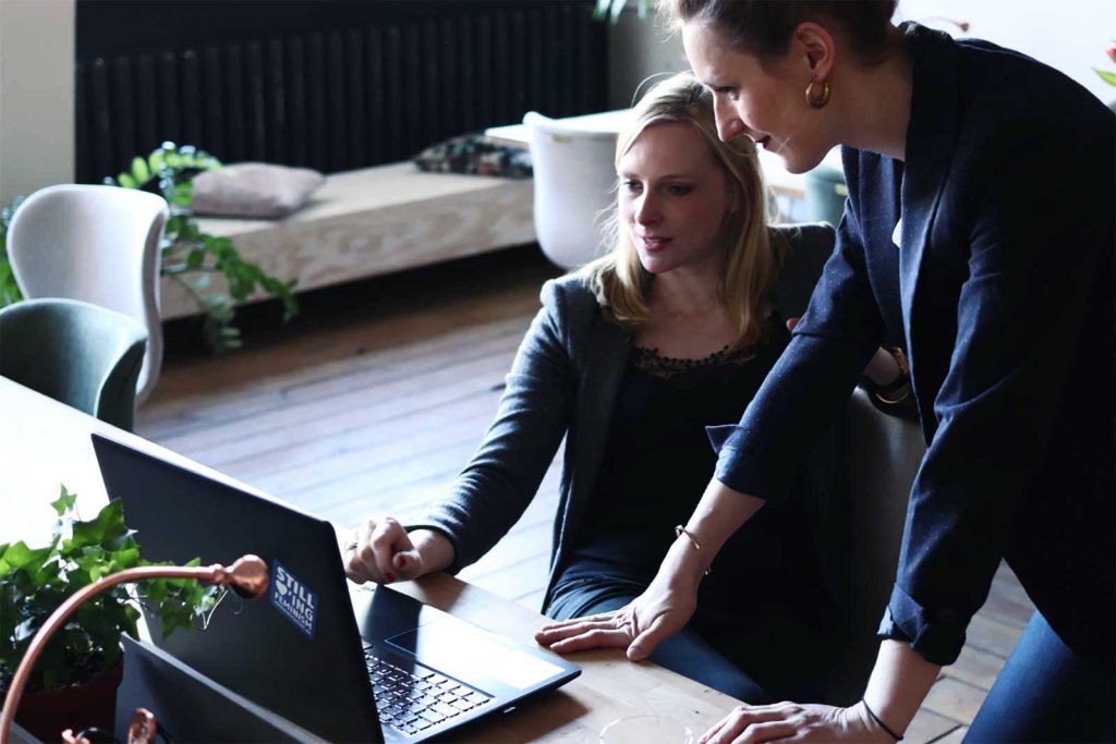 Two women are working together in an office setting. One is sitting at a table, using a laptop, while the other is standing, leaning over to point at something on the screen. There are plants and modern furniture in the background.