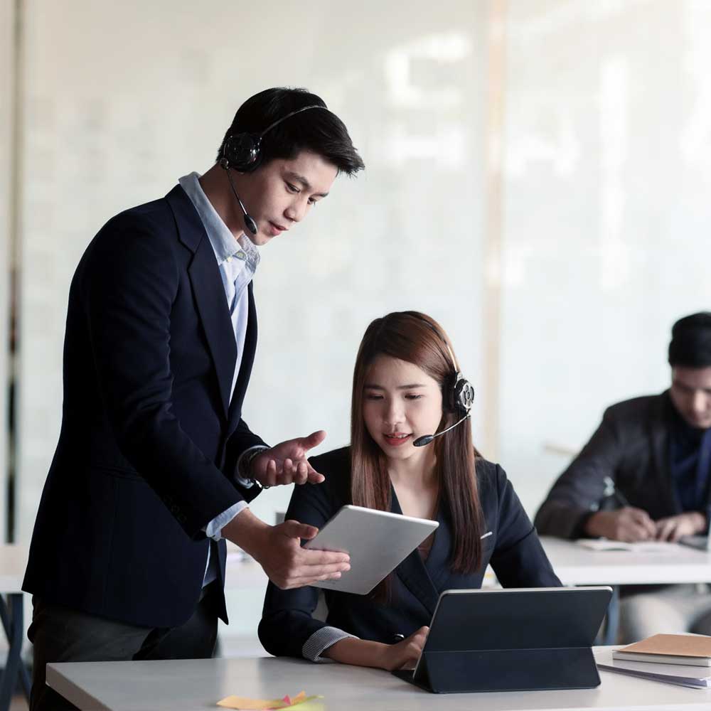 A man and a woman, both wearing headsets, are working together in an office. The man is holding a tablet and pointing at it while the woman looks at the screen attentively. Another person is visible in the background working on a laptop.