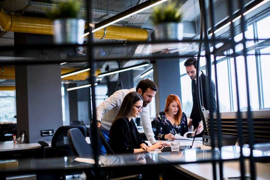 A group of four people in a modern office setting collaborate around a laptop. Two are standing and two are seated, engaged in a discussion. The room features industrial design elements and large windows letting in natural light.