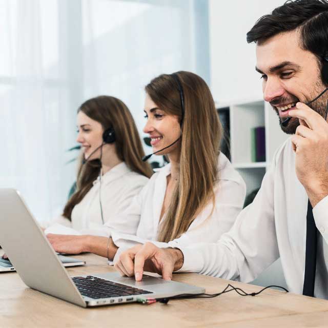 Three customer service representatives, wearing headsets and white shirts, sit at a desk working on laptops in an office. They are smiling and appear to be engaged in assisting customers. The background includes shelves and a window with natural light.