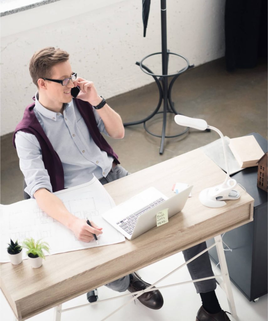 A person wearing glasses and a sweater over their shoulders is sitting at a desk, talking on the phone. The desk has a laptop, a blueprint, plants, and a lamp. The workspace appears modern and well-lit.