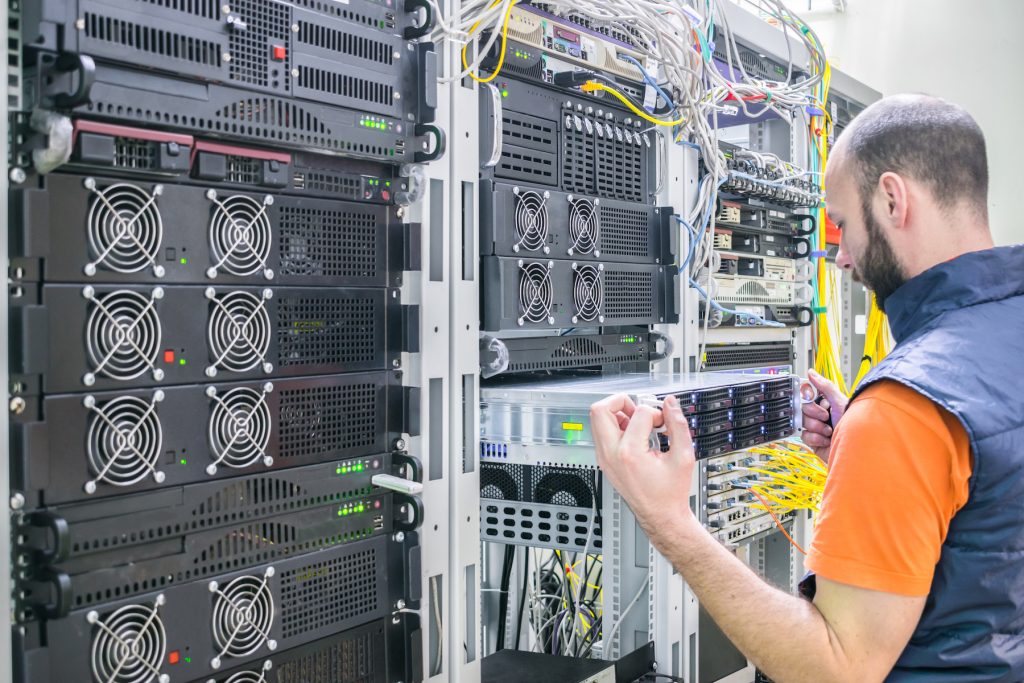 A man with short hair and a beard wearing an orange shirt and a dark vest is working in a server room. He is connecting cables to server racks filled with networking equipment and fiber optics, ensuring proper configuration in an IT services infrastructure setting.