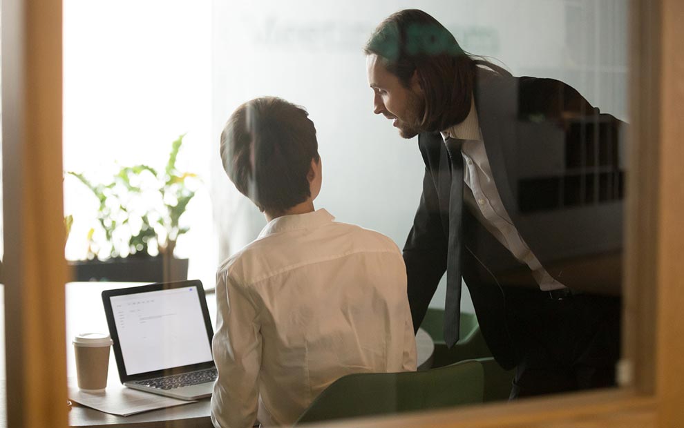 Two men in business attire are seen through a glass window. One man, sitting at a desk with an open laptop and a coffee cup, listens attentively to the other man, who is standing and leaning in, engaged in a serious conversation about IT services. Office plants are in the background.