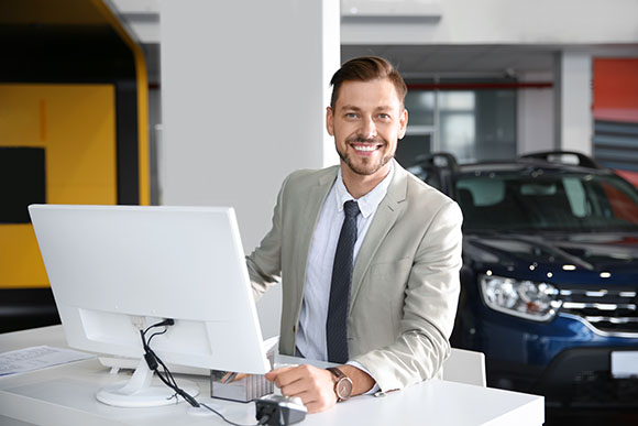 A smiling man in business attire sits at a white desk in front of a computer, seemingly providing managed IT services in an office or showroom with a dark-colored car in the background.