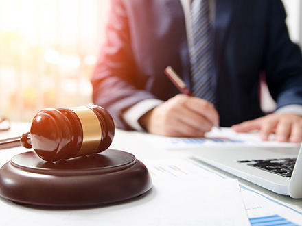 A wooden judge's gavel sits on a desk next to papers and a laptop. In the background, a person in a suit is visible, writing on a document. The scene, with its subtle hints of managed services and IT services, suggests a legal or courtroom setting intertwined with modern technology.