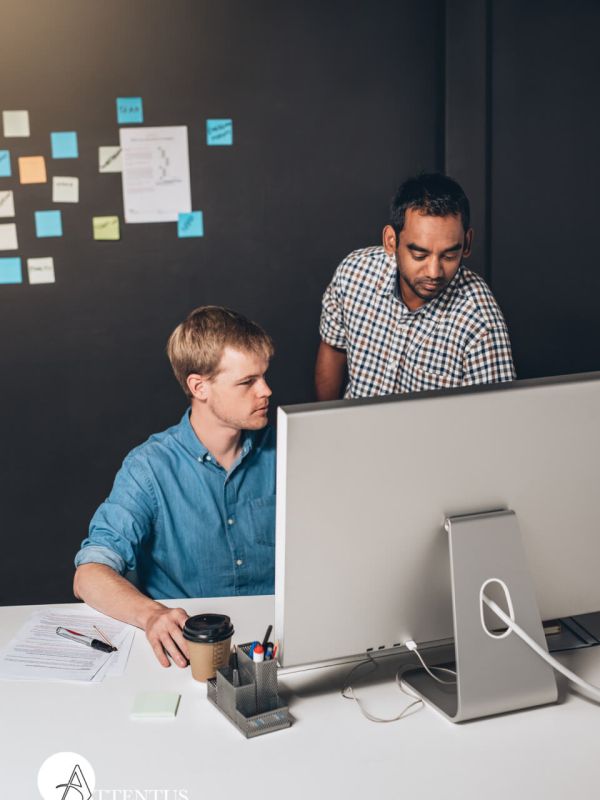 Two men collaborate at a desk with a large computer monitor in their Seattle office. One man sits, focused on the screen, holding a pen, while the other stands, pointing at the monitor. A coffee cup, papers, and office supplies are on the desk, and sticky notes are on the wall behind them—an IT consulting session in progress.